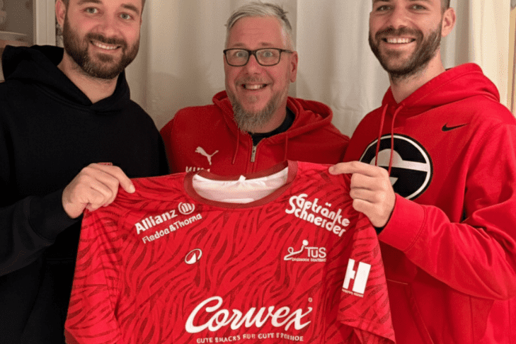 Drei Männer halten gemeinsam ein rotes Sporttrikot mit Sponsorenlogos vor einem weißen Vorhang.