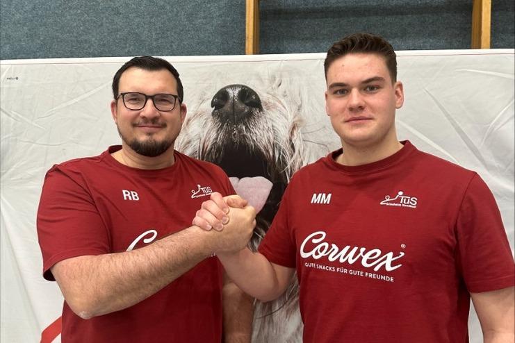 Zwei Männer in roten T-Shirts schütteln sich die Hände vor einem Plakat mit einem Hundekopf im Hintergrund.