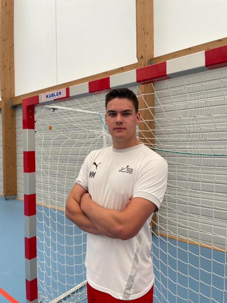 Junger Athlet steht mit verschränkten Armen vor einem Handballtor in einer Sporthalle.