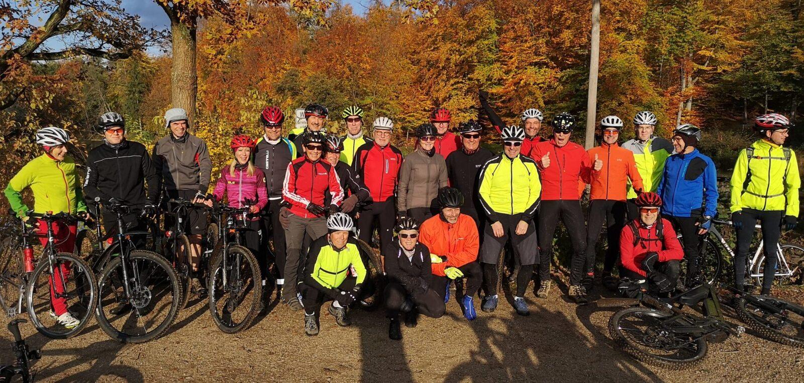 Gruppe von Radfahrern in bunter Kleidung posiert vor bunten Herbstbäumen. Fahrräder stehen daneben.