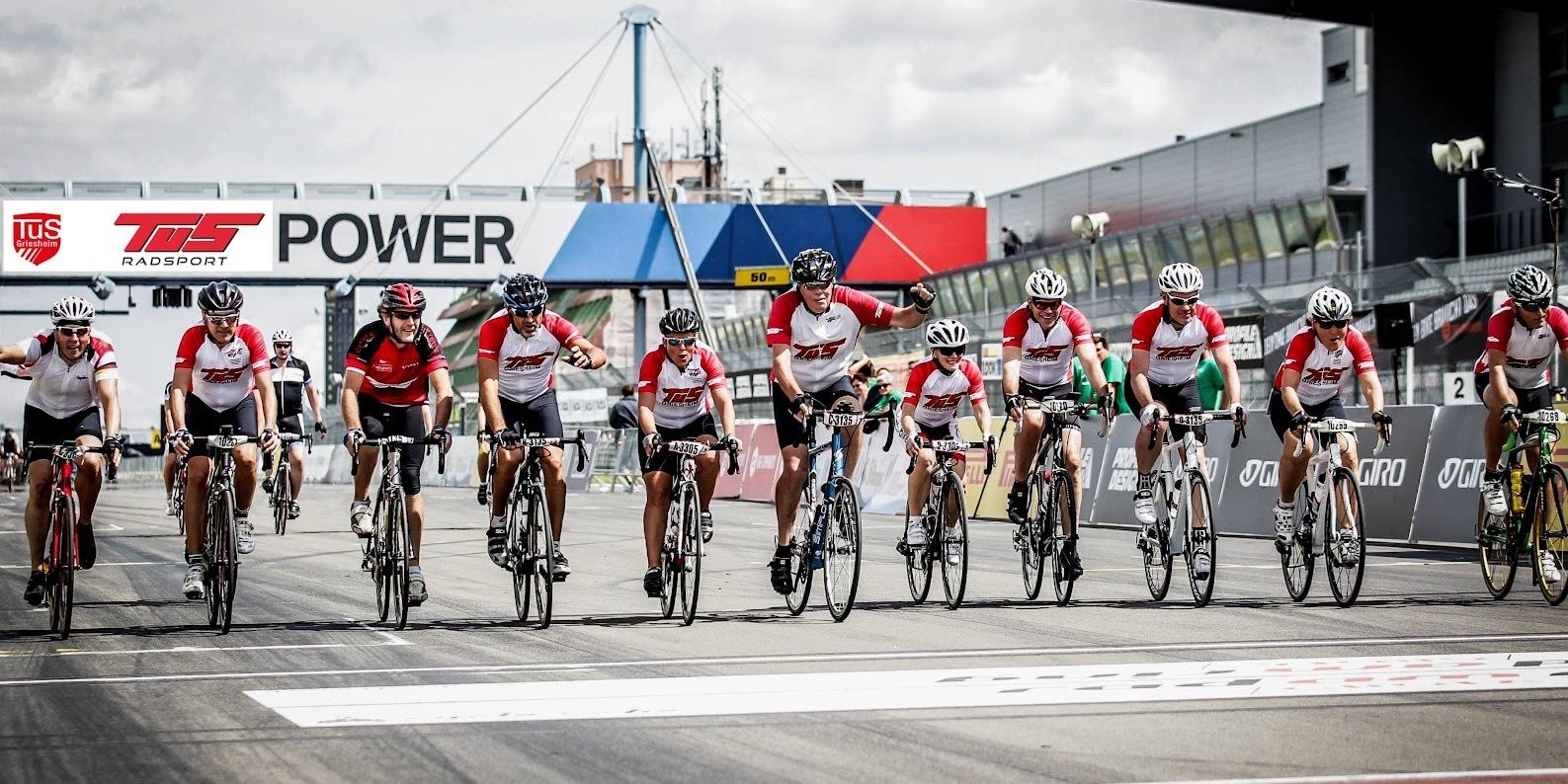 Gruppierung von Radfahrern in roten Shirts, die die Ziellinie auf einer Rennstrecke überqueren.