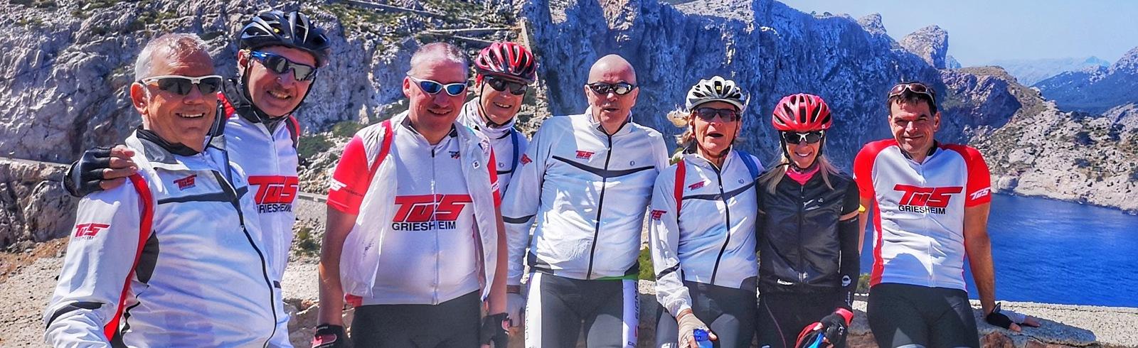 Gruppe von neun Radfahrern in Sportkleidung posiert vor einer Küstenlandschaft mit Bergen und blauem Wasser.