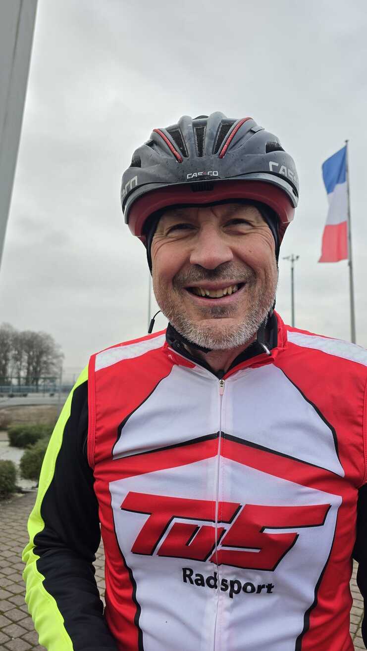 Ein radfahrender Mann mit Helm und roter Jacke steht lächelnd vor einem grauen Himmel und Fahnen.