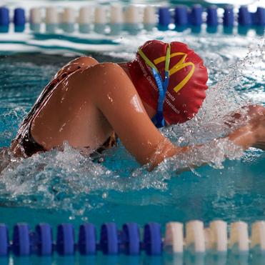 Schwimmerin in roter Badekappe vollzieht einen kraftvollen Rückenschwimmzug im Wasser, spritzt dabei Wasser auf.