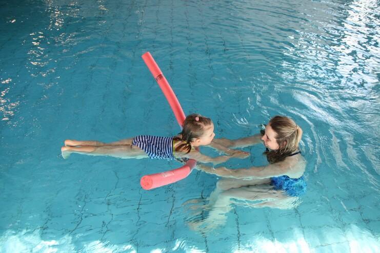 Zwei Mädchen im Wasser, das kleinere nutzt einen Schwimmnudel, während das andere sie hält und anlächelt.