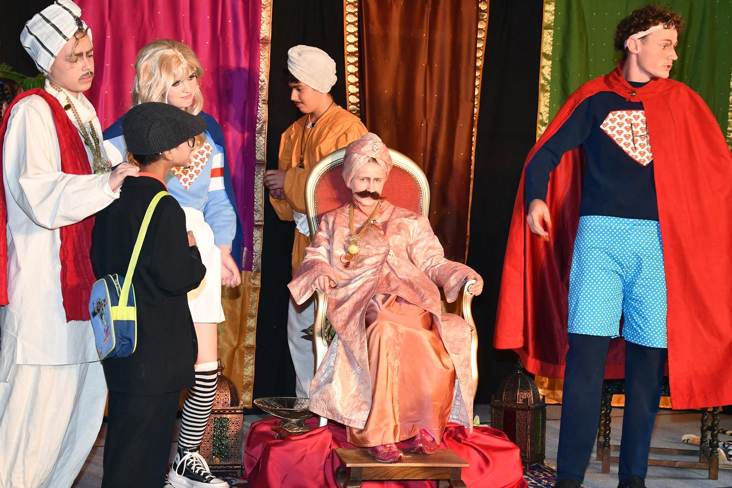 Szenenbild aus einem Theaterstück mit mehreren Kinderdarstellern in bunten Kostümen und einer sitzenden Figur in königlicher Kleidung.