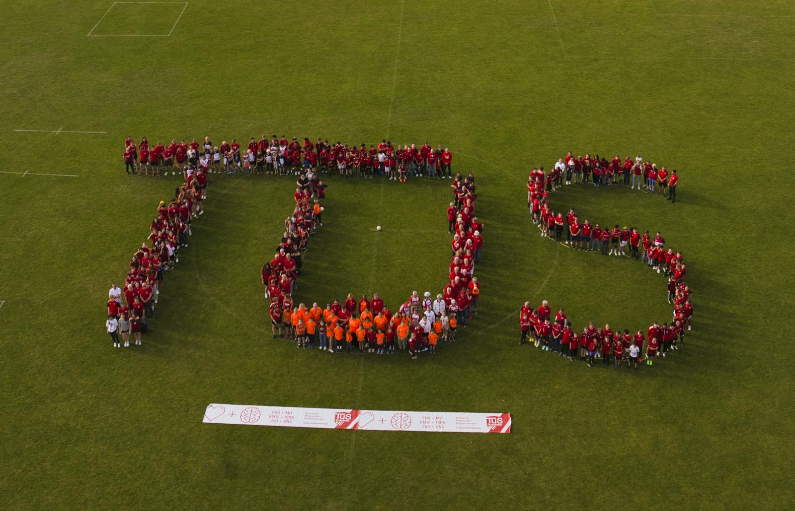 Gruppierung von Menschen in rot und orange bildet den Schriftzug "TDS" auf einem grünen Sportfeld unter blauem Himmel.