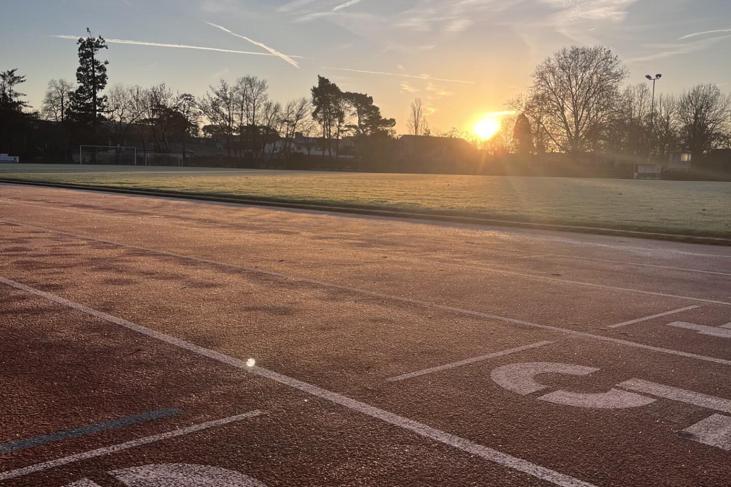 Sonnenaufgang über einer Laufbahn, mit Startlinien und frostbedecktem Gras im Hintergrund.