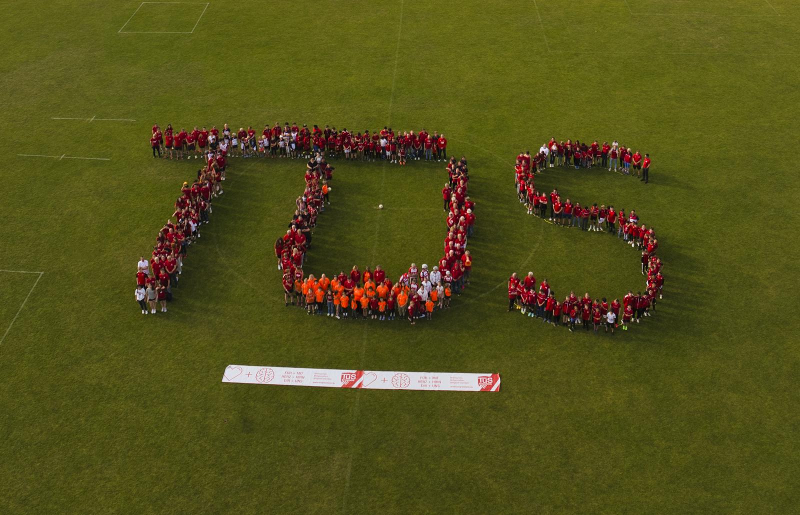 Gruppierung von Menschen in rot und orange bildet den Schriftzug "TDS" auf einem grünen Sportfeld unter blauem Himmel.